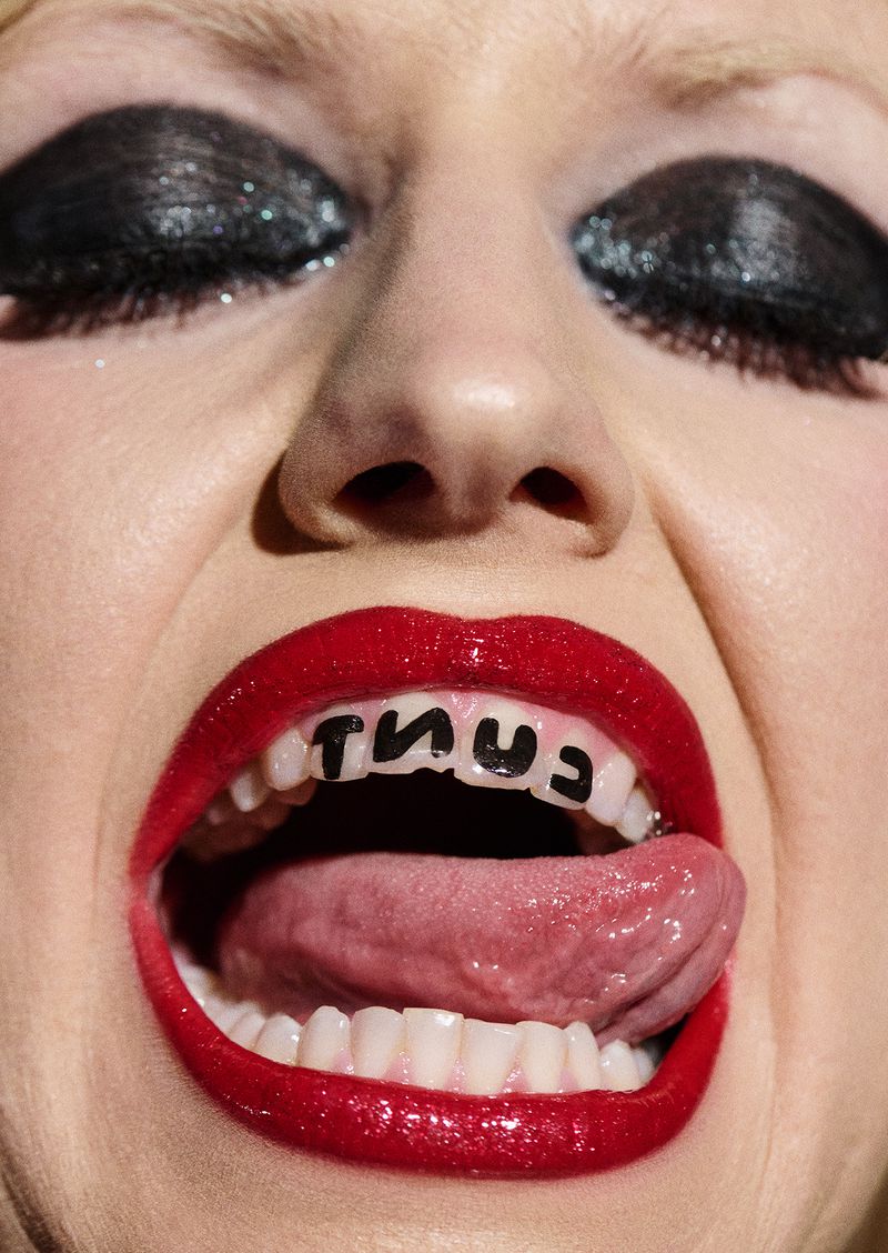Close-up shot of a woman's face with red lipstick and heavy black eye makeup, sticking out her tongue with text on her teeth.