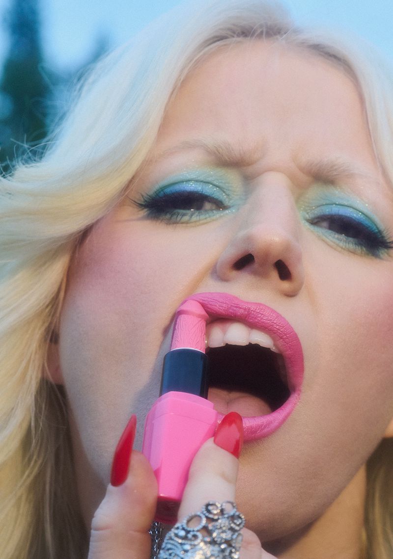 Close-up of a blond adult female applying bright pink lipstick, wearing long red nails and silver rings, against a neutral background.