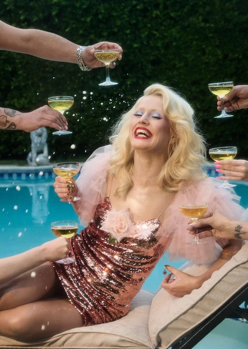 Blonde woman in a sparkling pink sequin mini dress laughing on a poolside lounger as hands toast her with champagne coupes.
