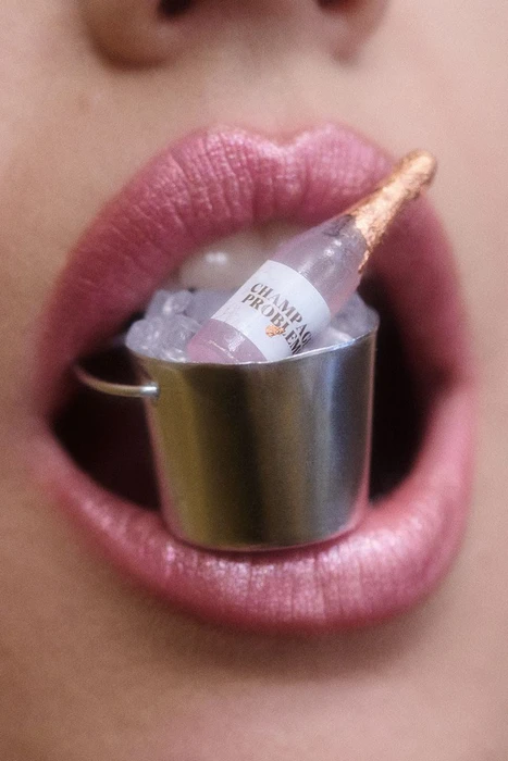 Extreme close-up of glossy pink lips holding a miniature champagne bottle inside a tiny metal ice bucket filled with ice cubes.