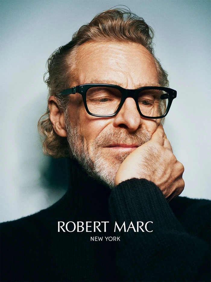 Man wearing thick black eyeglasses and a black turtleneck sweater against a light gradient background