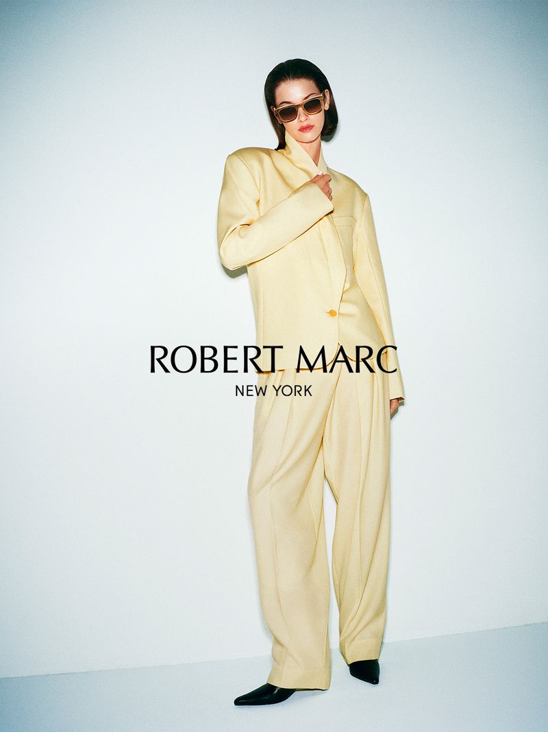 Model wearing yellow suit jacket, trousers, and sunglasses against a plain white studio backdrop with black typography