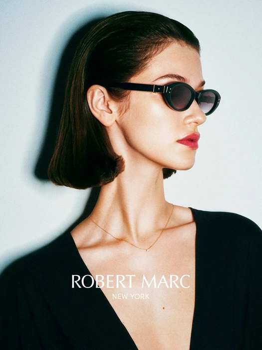 Model wearing dark sunglasses and black top against a pale background for the Robert Marc campaign shot by Guy Aroch.