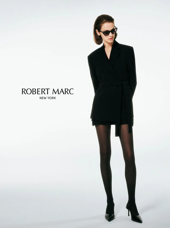 Model wearing dark sunglasses and a black belted blazer against a white background for Robert Marc
