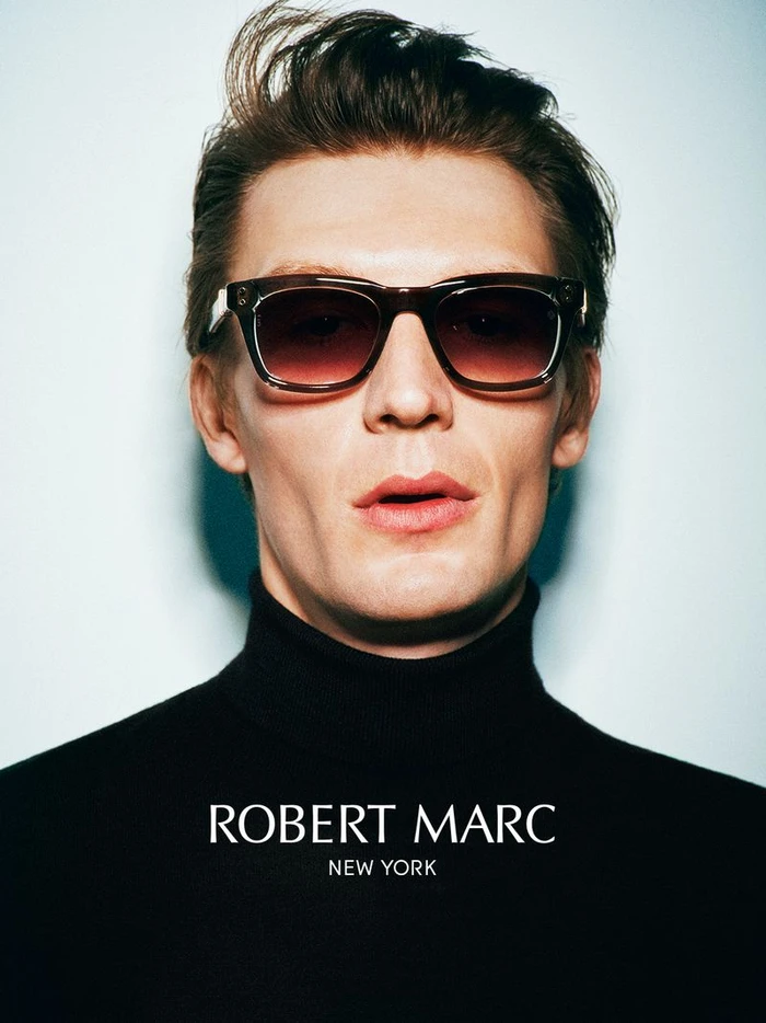 Close-up portrait of a man wearing dark tortoiseshell sunglasses and a black turtleneck against a light background