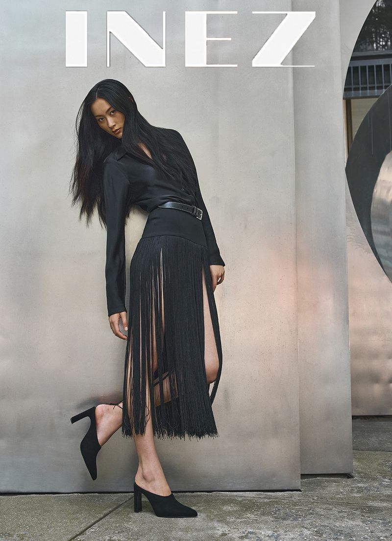 Model with long dark hair wearing a black fringe skirt and heeled mules leaning against a concrete wall outdoors.