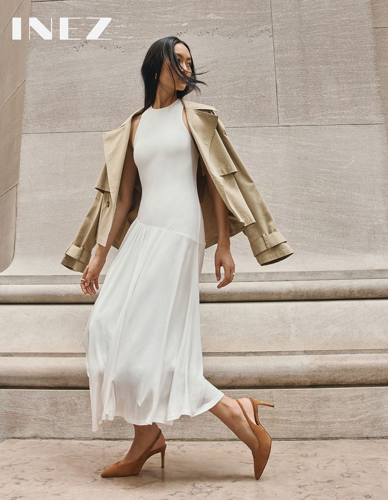 Model in a white midi dress and beige trench coat posing outdoors against monumental stone architecture