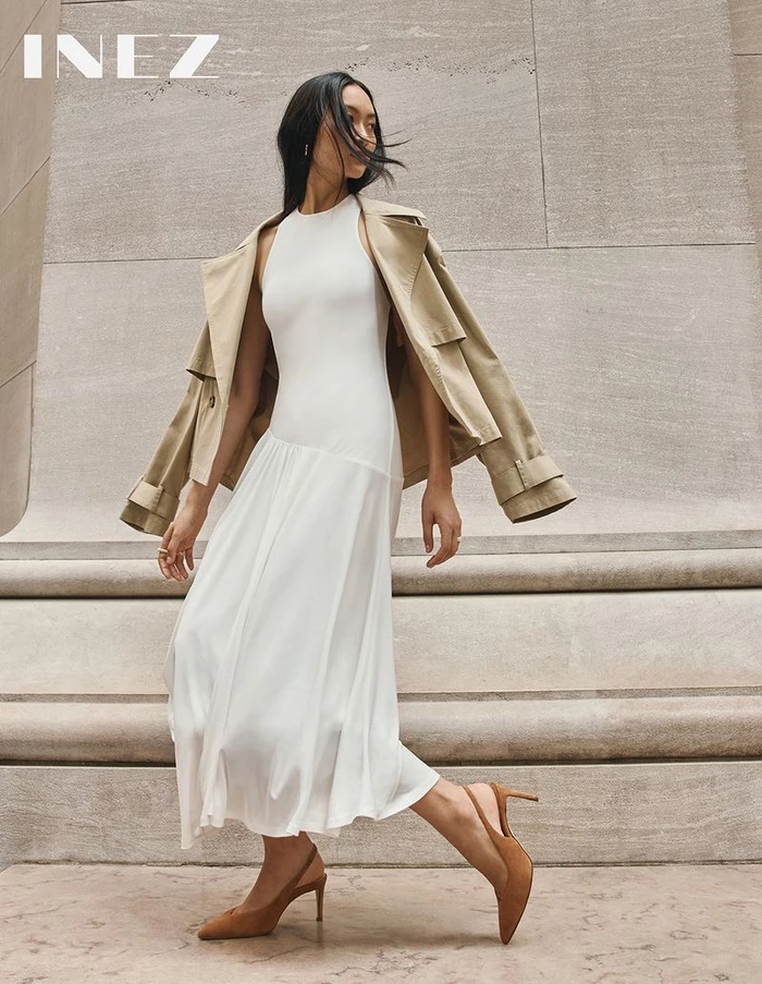 Model in a white midi dress and beige trench coat posing outdoors against monumental stone architecture