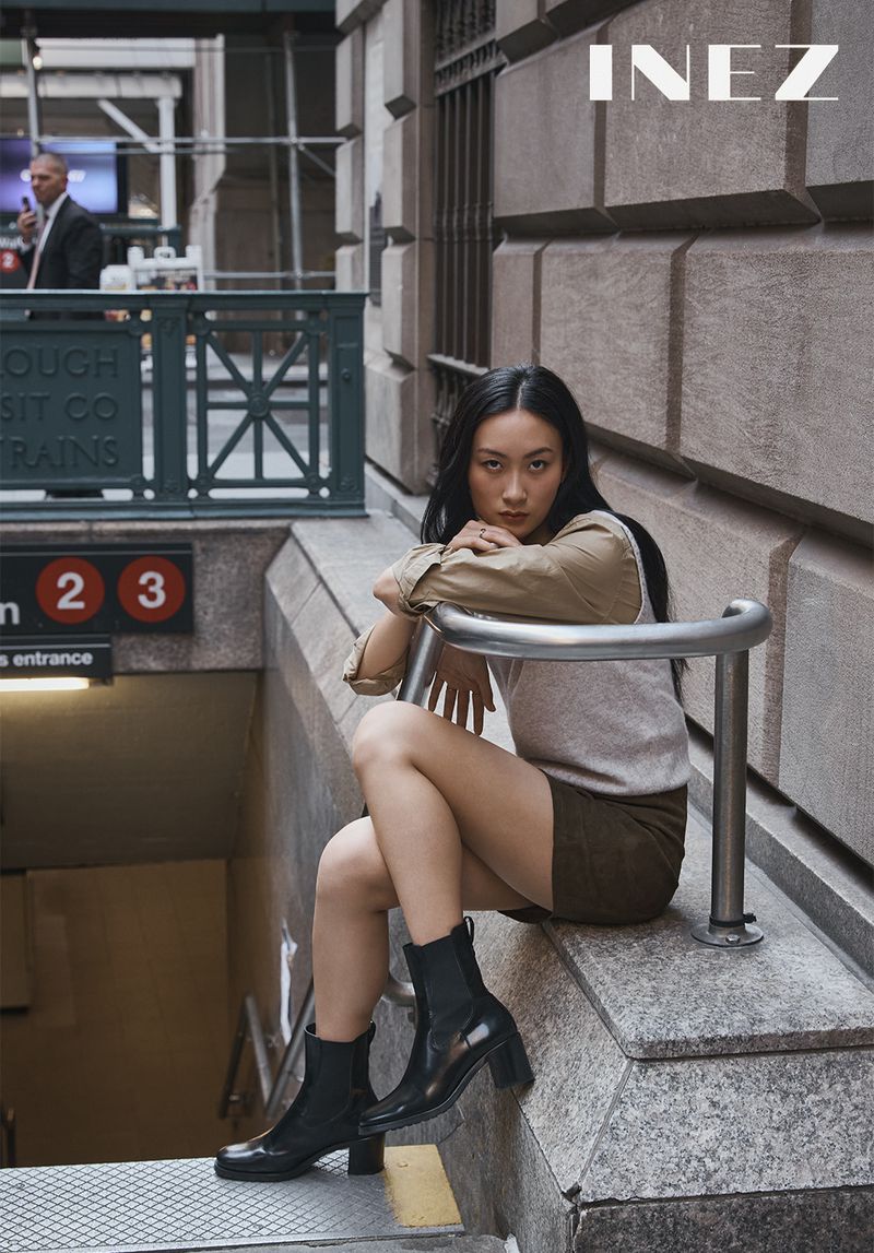Asian woman seated on a stone subway entrance railing wearing a tan shirt, knit vest, skirt, and black boots