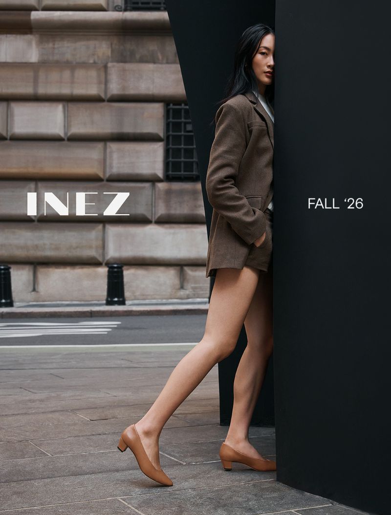 Woman in brown blazer and tan low-heel shoes leaning against a black structure outdoors in front of a stone building.