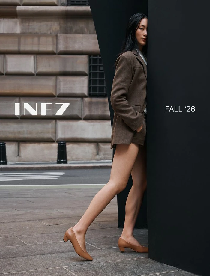 Woman in brown blazer and tan low-heel shoes leaning against a black structure outdoors in front of a stone building.