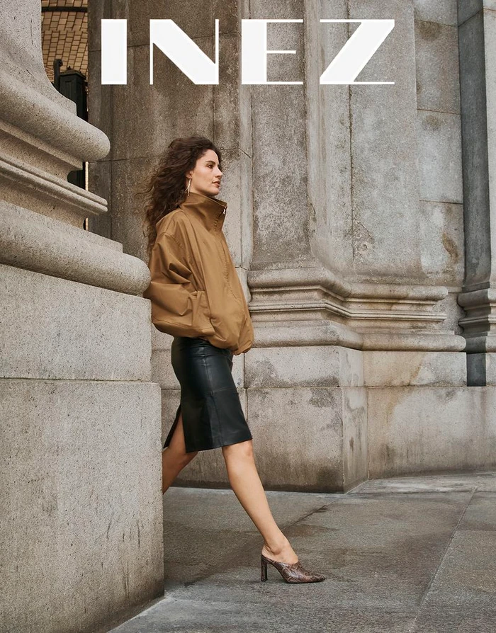 Woman with long curly hair wearing a brown jacket and black leather skirt walking past stone architectural columns