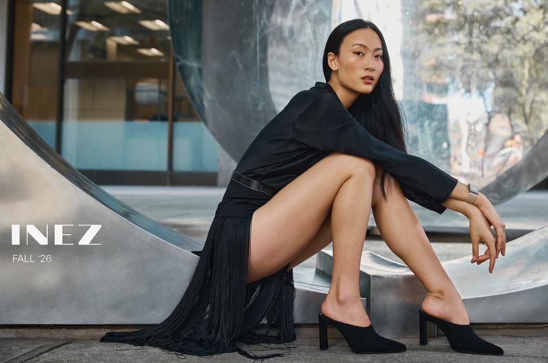 Asian woman with long dark hair wearing a black dress and heels seated outdoors near a large metallic sculpture