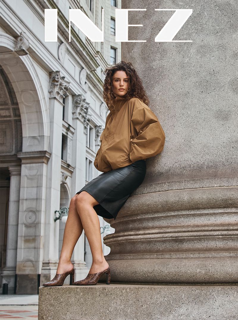 Woman with curly hair in a camel jacket and black skirt leaning against a large stone column on a city street