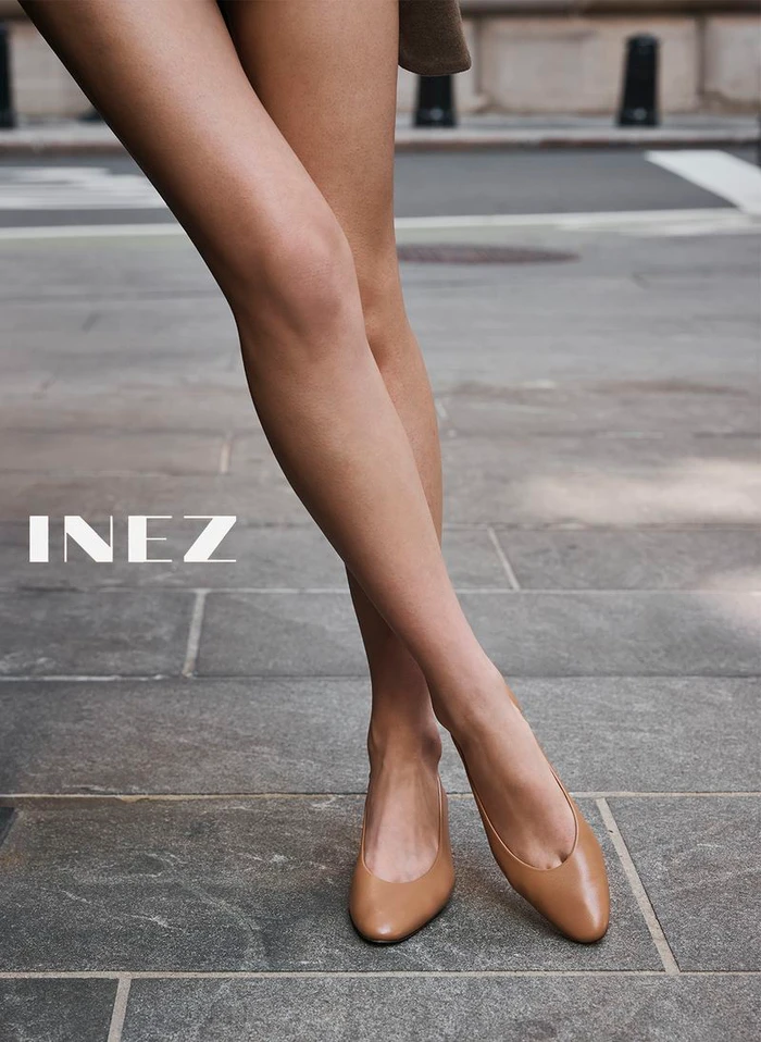 Close-up of a model standing on a stone sidewalk wearing tan leather flats for Inez Fall 2026 campaign.