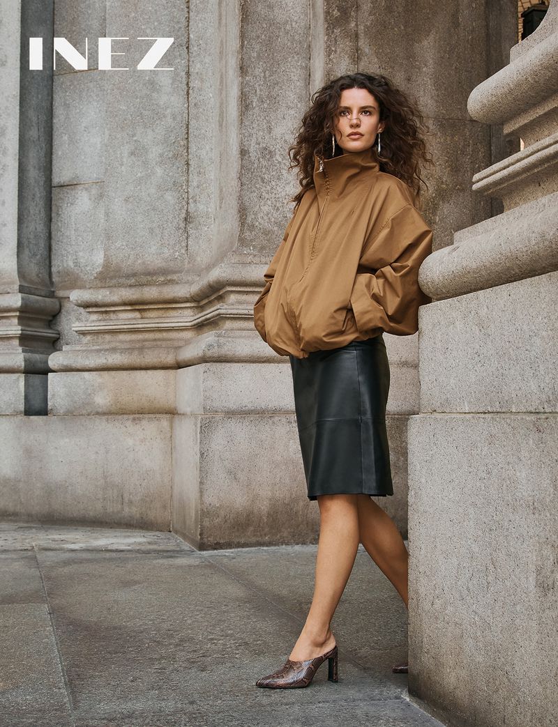 Woman with curly hair wearing a brown jacket and black skirt leaning against a stone building pillar.