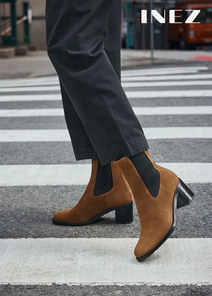 Close-up of a person wearing brown suede ankle boots and dark trousers standing on a city street crosswalk