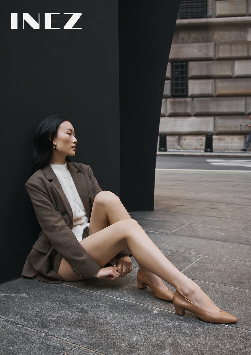 Model seated on stone pavement in a brown blazer and tan heels against a modern black wall