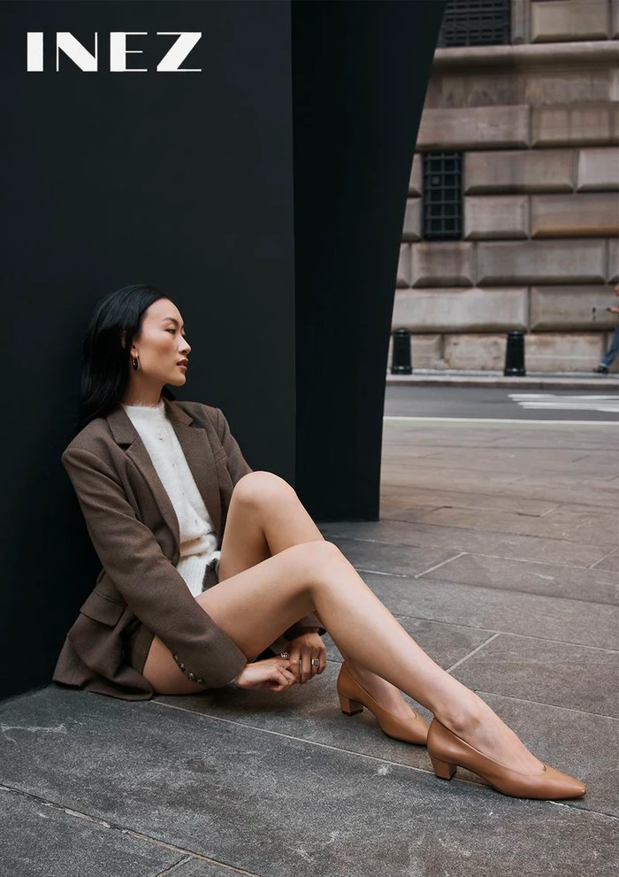 Model seated on stone pavement in a brown blazer and tan heels against a modern black wall