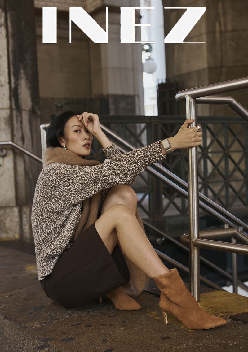 Woman wearing a patterned sweater and brown skirt sitting by a metal railing outdoors for Inez Fall 2026