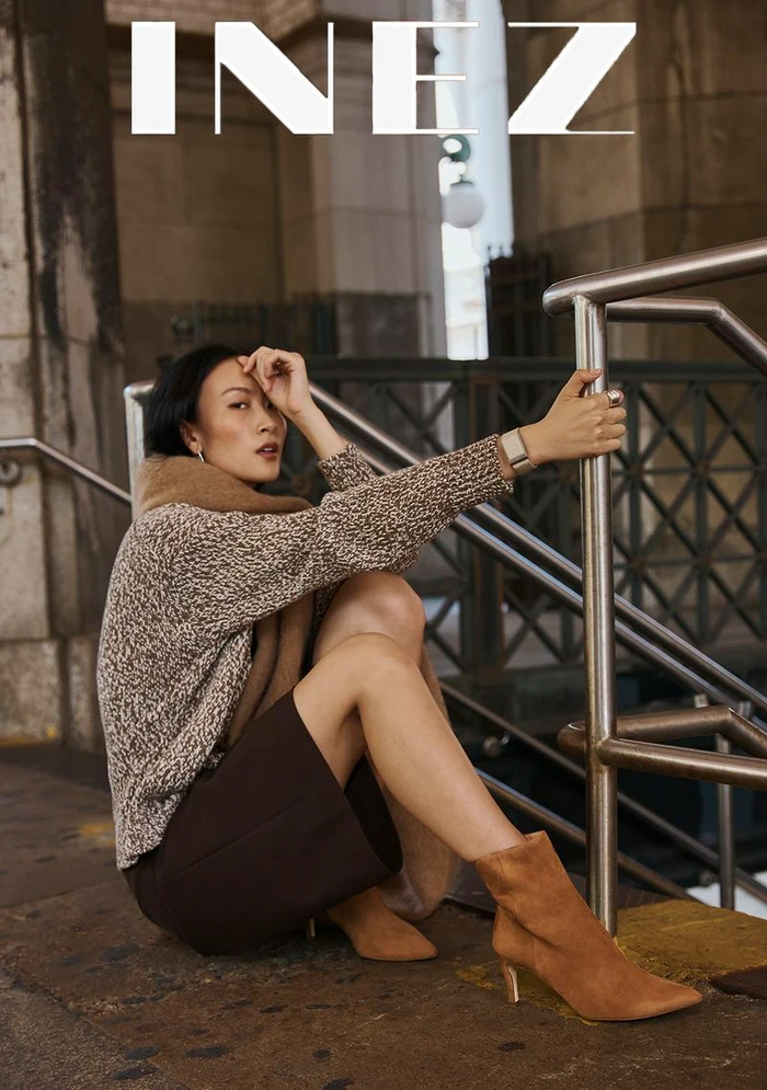 Woman wearing a patterned sweater and brown skirt sitting by a metal railing outdoors for Inez Fall 2026