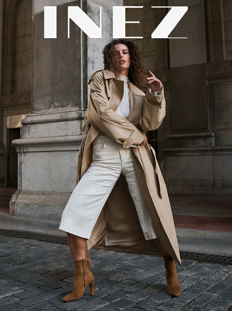 Model in a long beige trench coat and white trousers posing on stone pavement in front of classical architecture.