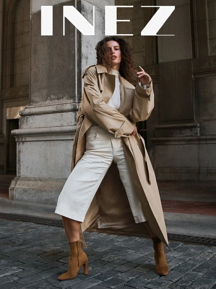 Model in a long beige trench coat and white trousers posing on stone pavement in front of classical architecture.