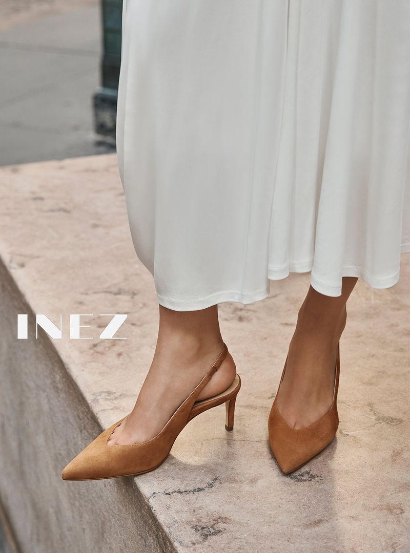 Close-up of a person wearing tan suede slingback kitten heels and a white midi skirt standing on a stone ledge