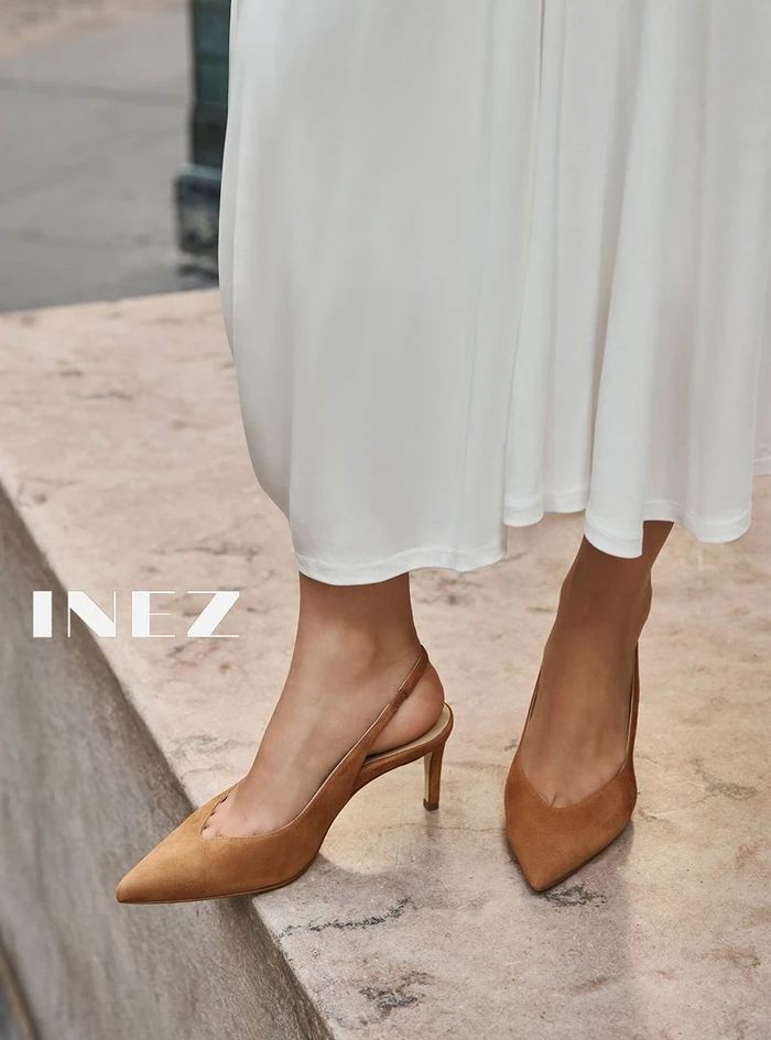Close-up of a person wearing tan suede slingback kitten heels and a white midi skirt standing on a stone ledge