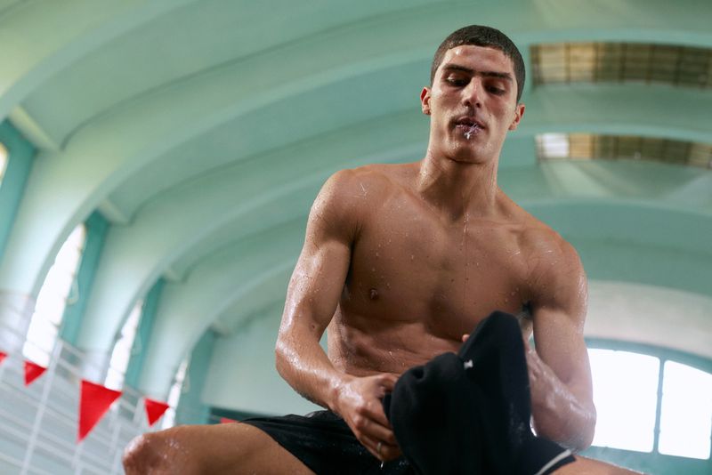 Sweaty shirtless man holding dark clothing inside an indoor swimming pool venue with arched ceilings.