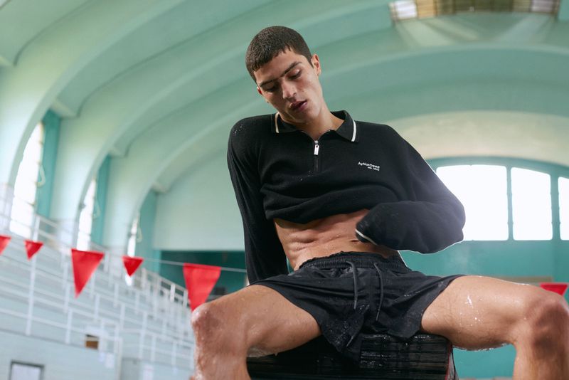 A young male model in black shorts and a black long-sleeve polo sitting indoors by an indoor swimming pool.