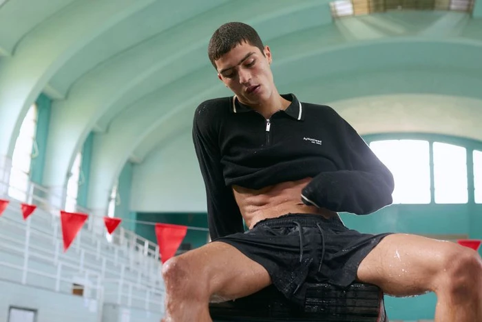 A young male model in black shorts and a black long-sleeve polo sitting indoors by an indoor swimming pool.