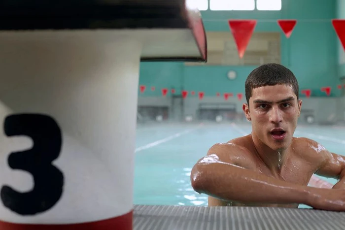 A shirtless male model leaning by a pool edge with a large number three block in the foreground for Ay Not Dead.