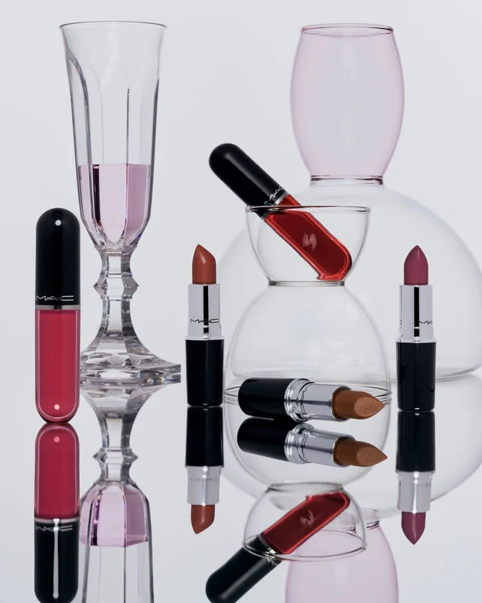 MAC Lustreglass lipsticks and glassware arranged on a reflective surface against a plain light background