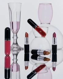 MAC Lustreglass lipsticks and glassware arranged on a reflective surface against a plain light background