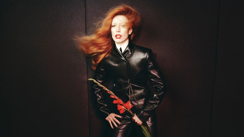 Raquel Zimmermann with windblown red hair wearing a black leather jacket holding red flowers for Khaite