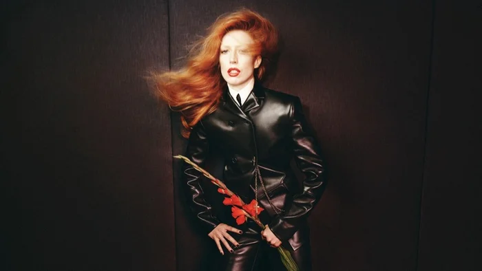 Raquel Zimmermann with windblown red hair wearing a black leather jacket holding red flowers for Khaite