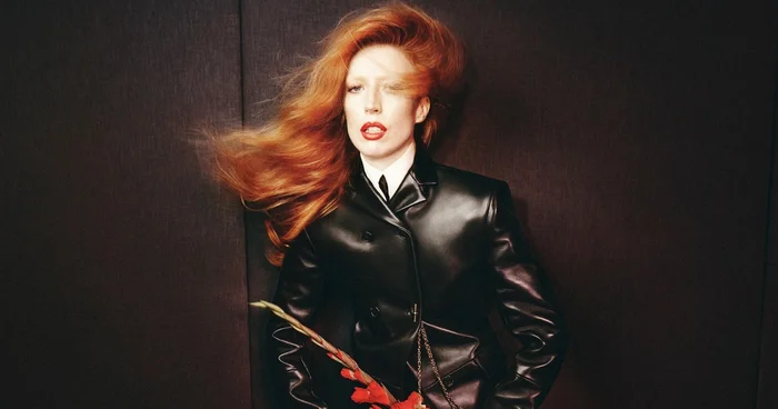 Model Raquel Zimmermann in a black leather jacket with long red hair against a dark textured background for Khaite Fall-Winter 2026 campaign.