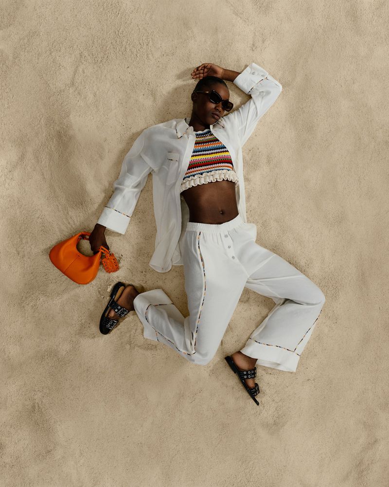 Woman in stylish white linen clothes poses on beige sand, wearing a colorful top, sunglasses and holding an orange handbag in studio.