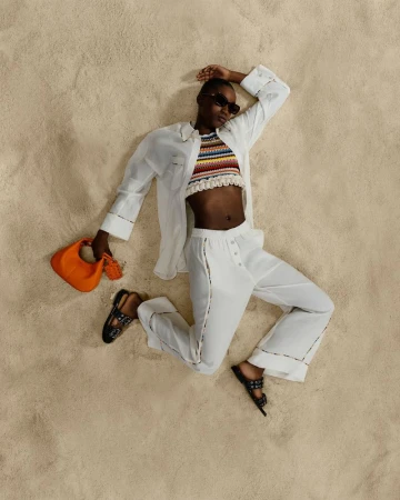 Woman in stylish white linen clothes poses on beige sand, wearing a colorful top, sunglasses and holding an orange handbag in studio.