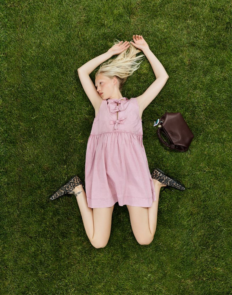Overhead shot of a fair-skinned woman lying on green grass in a pink dress with a brown bag next to her.