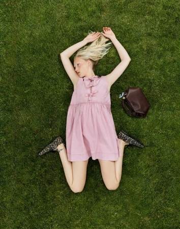 Overhead shot of a fair-skinned woman lying on green grass in a pink dress with a brown bag next to her.