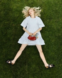 Blonde model lying on green grass wearing a striped dress, black studded sandals, and holding a red handbag