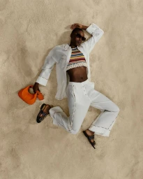 Black woman in a white linen shirt, crochet crop top, and white trousers lying on sand holding an orange handbag.