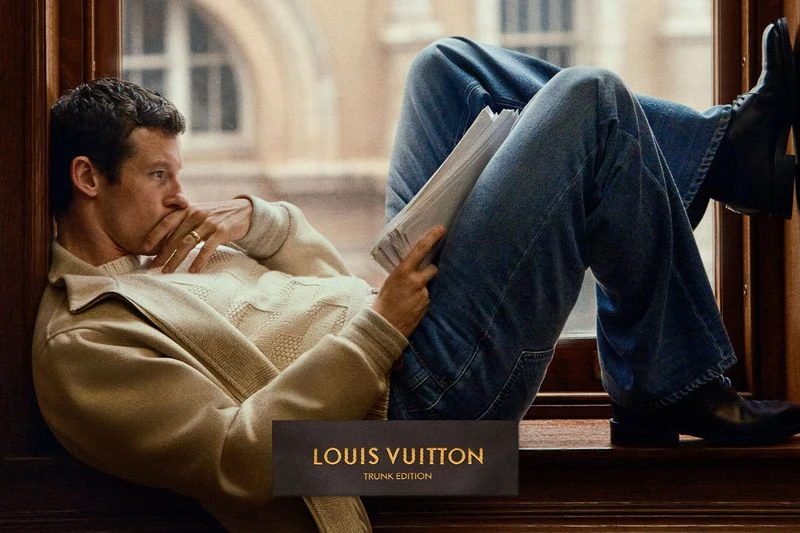 Callum Turner in a cream cardigan and jeans lounging by a wooden window for Louis Vuitton