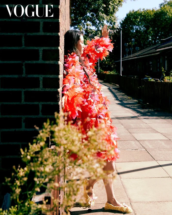 Model wearing a vibrant red and pink textured garment leaning against a brick wall on a sunlit street.