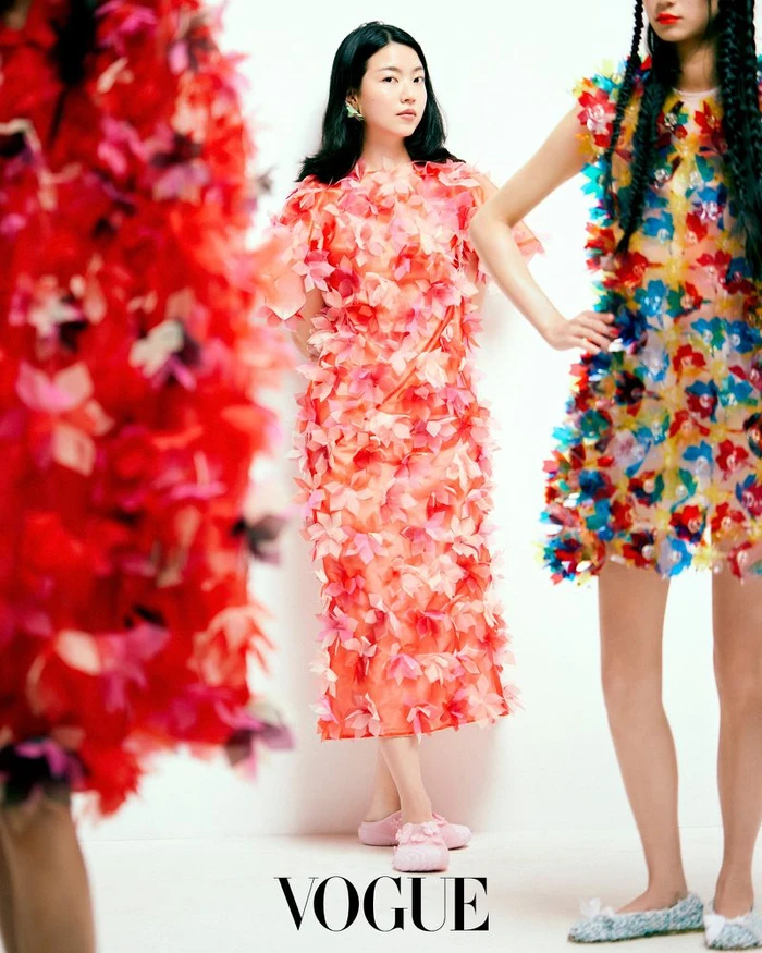 Model standing against a white wall wearing a pink floral textured dress and pink shoes, with other figures in the foreground and beside her.