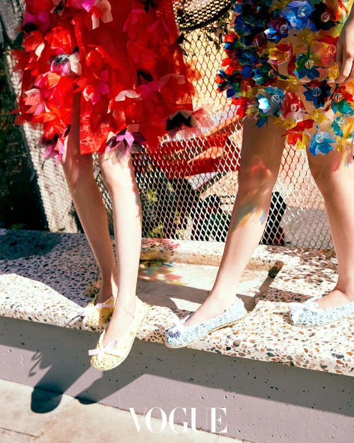 Two models wearing decorative colourful skirts and flat shoes standing on a low stone wall outdoors for Vogue Korea.