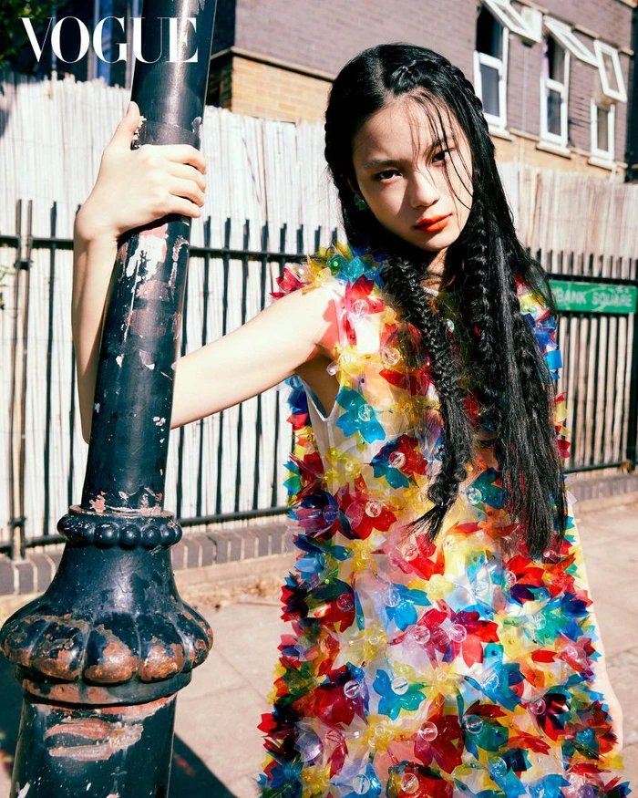 A woman in a colourful floral embellished dress leaning against a dark post on a city street.