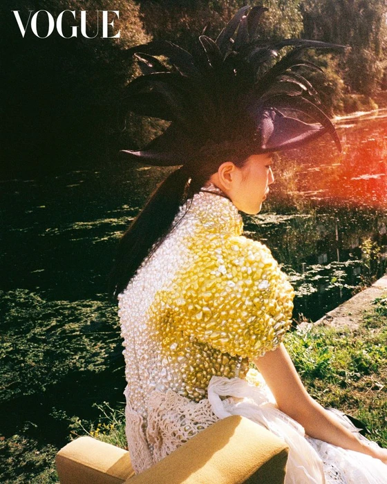 Model wearing an elaborate feathered hat and heavily embellished pearl dress seated near water for Vogue Korea.